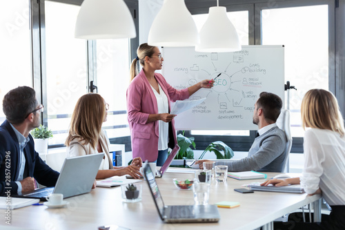 Fotografía Elegant young businesswoman pointing at white blackboard and explain a project to her colleagues on coworking place