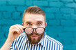 © satura_ - Close-up portrait of a curious surprised young man in glasses with a mustache and beard posing on a background of blue blurred brick wall. Concept of surprise and shocking information.