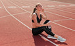 © splitov27 - Slim Fit woman in sportwear listening to music with earphones while sitting on a stadium track with a red coating in the open air. Running, healthy lifestyle concept