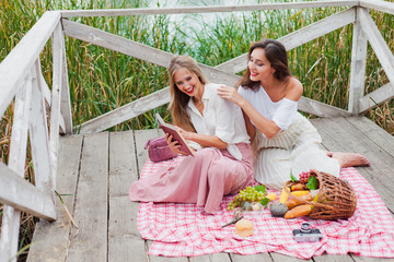 Two cheerful young women have a picnic outdoors on a summer day. Two girlfriends in retro vintage style clothes spend time together on the pond pier.