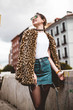 Stylish portrait of young woman looking aside smiling and wearing trendy animal, leopard print faux fur coat, fashion sunglasses, posing in Madrid city