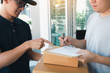 © wutzkoh - Delivery young man standing at the door of home and carrying parcels for young male to signing.