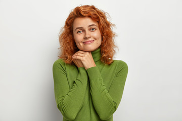 Lovely redhead woman keeps hands together near face, looks tenderly at camera, smiles gently, wears green jumper, poses against white background, has healthy skin, no make up. Facial expressions