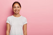 Studio shot of happy young Asian woman has tender smile, looks aside with charming expression, wears casual white t shirt, has natural beauty, isolated on pink wall. People and emotions concept