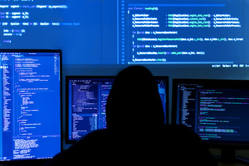 Back view of man in black hood is hacking password. Programmer is writing malware on multiple screens to steal private data. Program code from projector light on wall. Hacker attack concept.