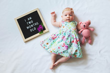 Two Months Old Baby Girl Wearing Floral Dress And Headband Laying Near Text Board And Hugging Teddy Bear.