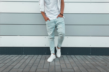 Wall Mural - Young stylish man in a white fashionable t-shirt in vintage blue jeans in white trendy sneakers stands near a modern wall. Fashionable men's summer clothing. Street style. Close-up of the male body.