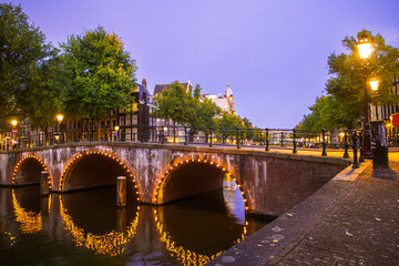 Fototapete - Amsterdam seen in the evening with light, canal and bridge