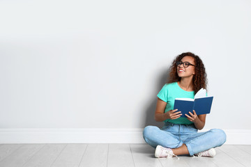 Beautiful young African-American woman reading book on wooden floor near light wall, space for text