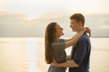 Teenagers look at each other on beach, cloudy weather and sunset on background