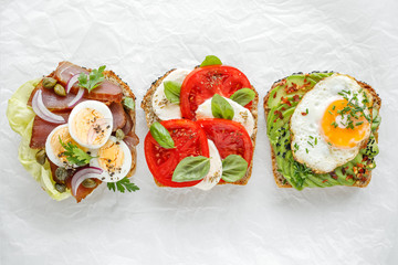 Wall Mural - Variety of open sandwich,  wholemeal bread slices  with various toppings: smoked tuna, egg, lettuce, tomatoes, mozzarella, avocado, fried egg, fresh herbs on a white background, top view. 