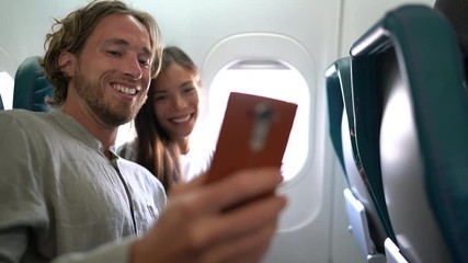 Wall Mural - Couple in plane on travel taking selfie photo or video while flying on vacation travel. Two happy millennials taking pictures for social media going on honeymoon.