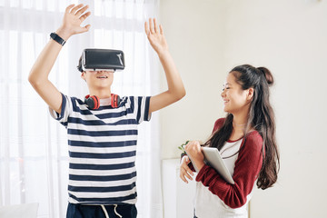 Wall Mural - smiling mixed race teenage girl looking at her brother playing video game in virual reality headset
