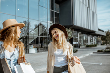 Wall Mural - Two happy girlfriends walking with shopping bags in front of the shopping mall, feeling satisfied with new purchases