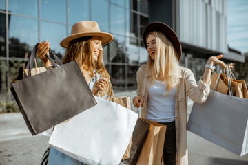 Two happy girlfriends walking with shopping bags in front of the shopping mall, feeling satisfied with new purchases