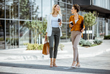 Full Body Portrait Of A Two Young Businesswomen Walking With Coffee Cups Near The Modern Office Building Outdoors
