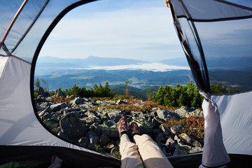 Wall Mural - Camping on the top of mountain on summer morning. Young hiker woman sitting in the entrance of tourist tent. On foggy mountains background. View of legs. Point of view shot. Tourism adventure concept