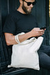 © 4Max - Young man holding white textile eco bag against urban city background. . Ecology or environment protection concept. White eco bag for mock up.