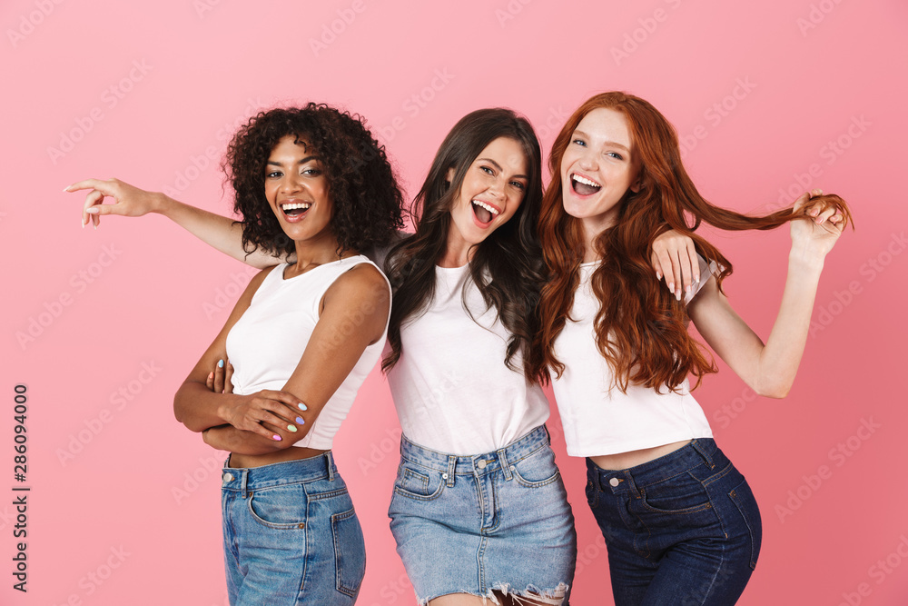 Cheery young three multiethnic girls friends posing isolated over pink wall background. - obrazy, fototapety, plakaty 