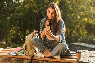 Wall Mural - Image of attractive brunette woman using smartphone and earphones while sitting at bench in green park