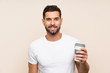 © luismolinero - Young man with beard holding a take away coffee over isolated blue background smiling a lot