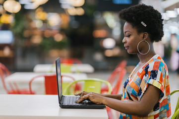 Young african american girl sitting at cafe table with laptop and looking at screen. Dark skinned freelancer concept.