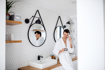 Young man with coffee and smartphone in the bedroom, making a phone call.