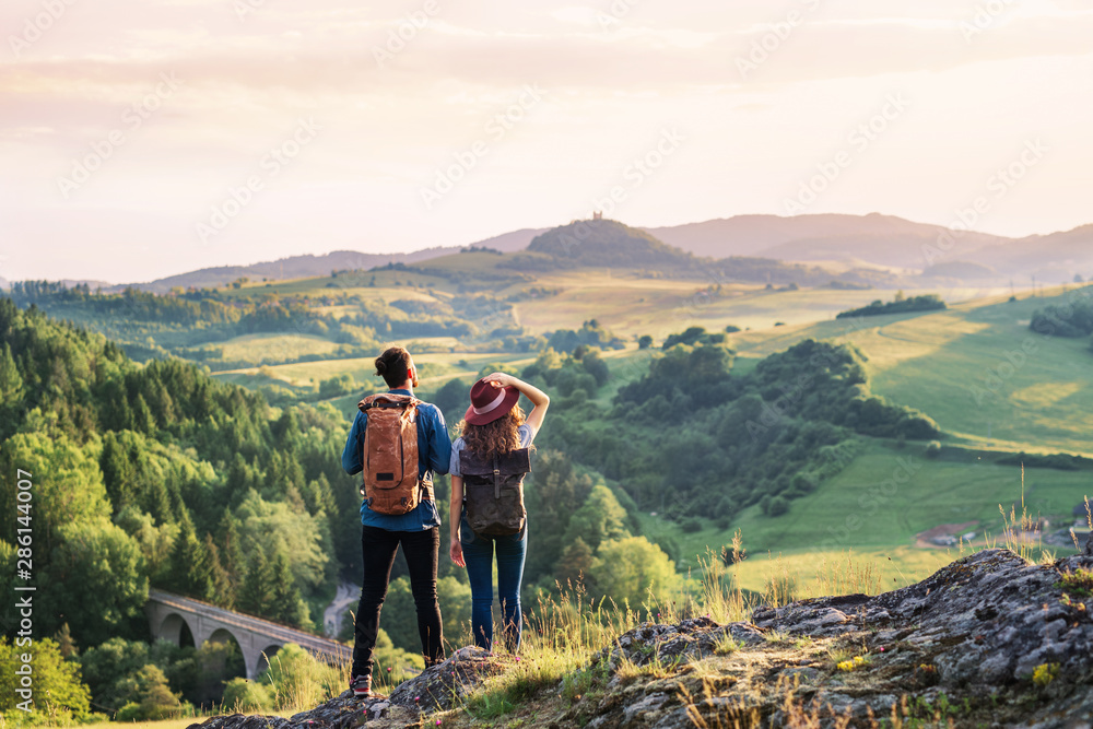 Rear view of young tourist couple travellers hiking in nature, resting. - obrazy, fototapety, plakaty 