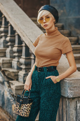 Street fashion portrait of young elegant woman wearing leather beret, beige glasses, turtleneck, green corduroy high waist trousers, golden wrist watch, holding trendy leopard printed box bag