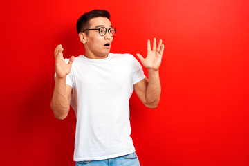 Shocked Asian man dressed in white casual and glasses with open mouth and hands up looks to the side. Surprised young Kazakh student guy on red studio background