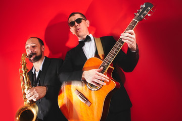 Group of two musicians, male jazz band, guitarist and saxophonist in classical costumes improvise on musical instruments in a studio on  red background