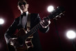 © amixstudio - Portrait of classical musician with guitar in red studio with stage lighting. Guitarist in black glasses and suit with a bow tie improvises on instrument