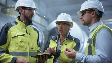 Wall Mural - Three Heavy Industry Engineers Stand in Pipe Manufacturing Factory, Use Digital Tablet Computer, Have Discussion. Large Pipe Assembled. Design and Construction of Oil, Gas and Fuels Transport Pipeline