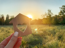 Miniature Wooden House In The Shape Of A Heart In The Hands Of A Girl Outdoors. Real Estate Concept. Eco-friendly Home. Buying A Housing Outside The City. Nature. Fresh Air. Sunset Light