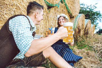 Poster - Junge Frau und mann mit Bierkrug und Brezel  im feld vor einem strohballen trinken bayerisches bier .Oktoberfest-Konzept 