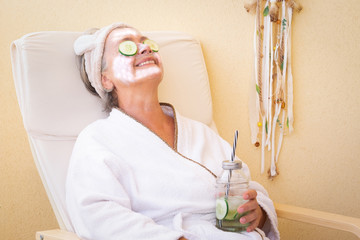 An elderly woman takes care of her face with a special cucumber-based cleaning mask. Sitting and relaxing outdoors on the terrace with a healthy drink. Bright light