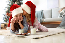 Gossiping Little Girls In Santa Claus Hats Eating Cookies With Milk At Home