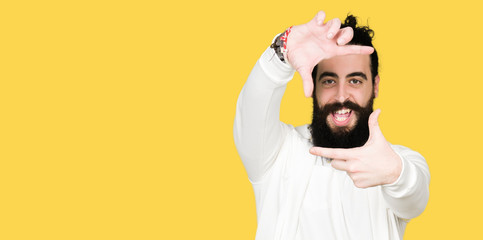 Wall Mural - Young man with long hair and beard wearing sporty sweatshirt smiling making frame with hands and fingers with happy face. Creativity and photography concept.