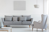 Fototapeta  - Big comfortable wing back armchair next to long grey scandinavian sofa with pillows in bright living room interior