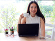 © Phuttharak - Asian woman holding cup of coffee smiling and looking to the camera.computer and glass and flower pot on table.