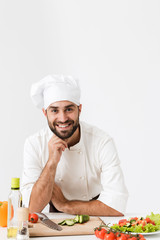 Wall Mural - Pleased positive happy young chef isolated over white wall background in uniform cooking with fresh vegetables.