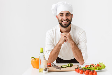 Wall Mural - Pleased positive happy young chef isolated over white wall background in uniform cooking with fresh vegetables.