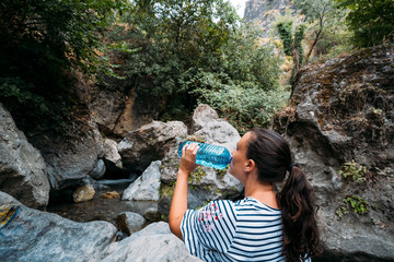 Young woman drinking water from plastic bottle and enjoying mountain sunset.