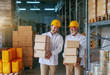 © Dusan Petkovic - Three quarter length of two smiling Caucasian storage employees in white uniforms and with yellow helmets carrying boxes in storage.