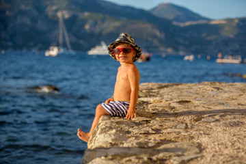 Wall Mural - Sweet toddler boy with summer hat and sunglasses, sitting on the beach shore on sunset, enjoying