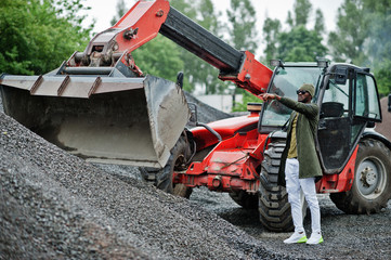 Stylish african american man in hat and sunglasses posed outdoor in rain against tractor with a bucket.