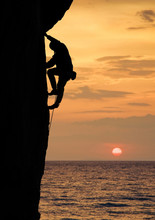 Silhouette Of Male Climber Ascenting High Up On Straight Cliff At Sunset. Side View. Dangerous And Risky Rock Climbing Above Ocean. Big Round Sun On Beautiful Orange Sky On Background. Copy Space