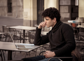 Attractive young man studying or working in a coffee shop with laptop in european city outdoors