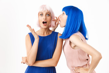 Portrait of two beautiful women wearing blue and pink wigs talking secrets in ear and expressing surprise