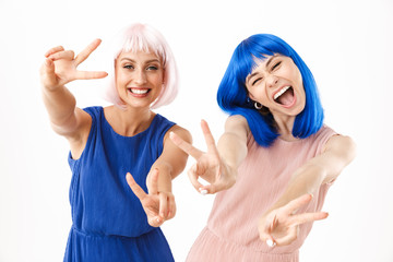 Wall Mural - Portrait of two excited women wearing blue and pink wigs smiling while gesturing peace sign